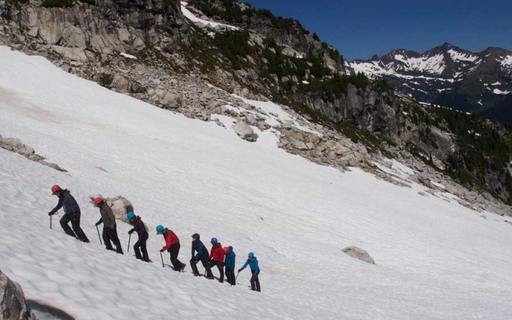 A group of people wearing mountaineering gear hike in a line up a snowy incline